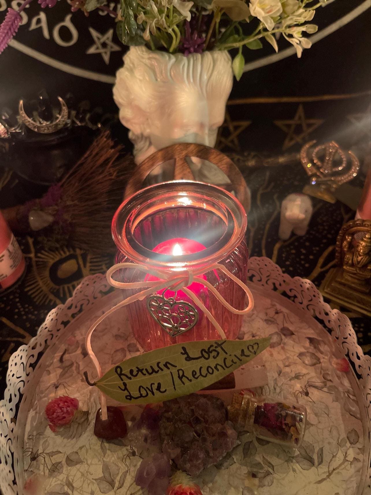 May include: A pink glass candle holder with a lit candle inside. The candle holder is on a white lace doily with a pink crystal, a small glass bottle with dried flowers, and a leaf with the text 'Return Lost Love/Reconcile' written on it.