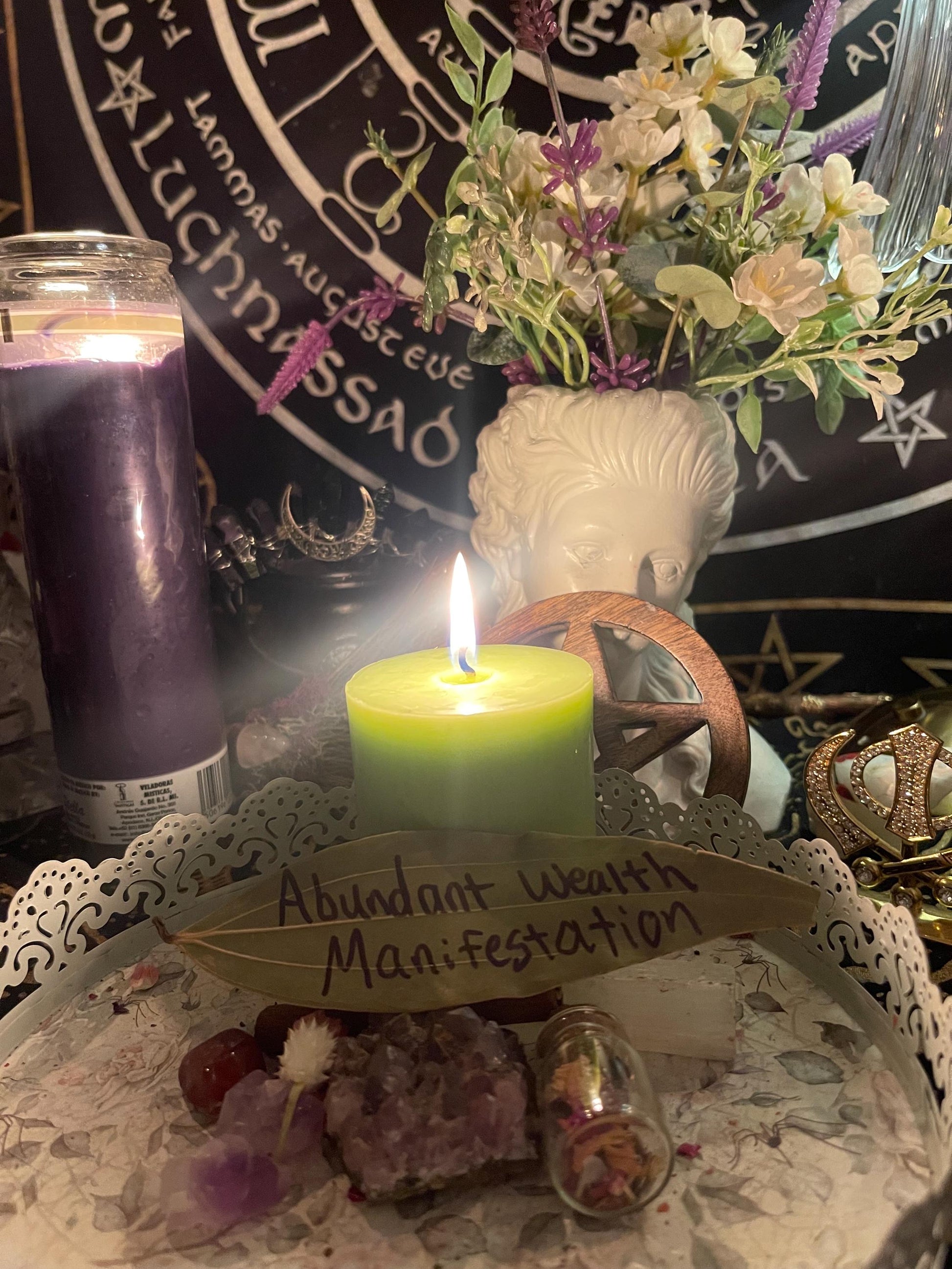 May include: A green candle with a flame on top, a small glass bottle with dried flowers inside, and a purple crystal on a white lace doily. The doily is on a black background with a pentagram and the words 'Abundant Wealth Manifestation' written on a leaf.